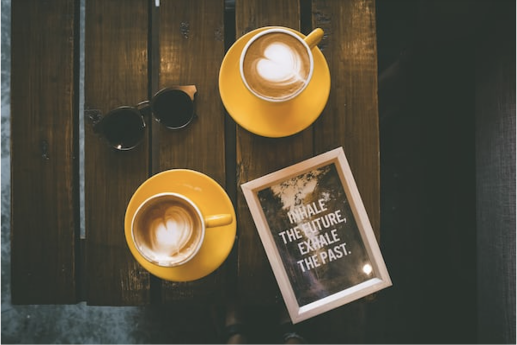 In this bird's eye image, a pair of sunglasses, two latte's in yellow mugs and a picture frame tat reads " Inhale the future, Exhale the past" sits on a dark wood surface. 