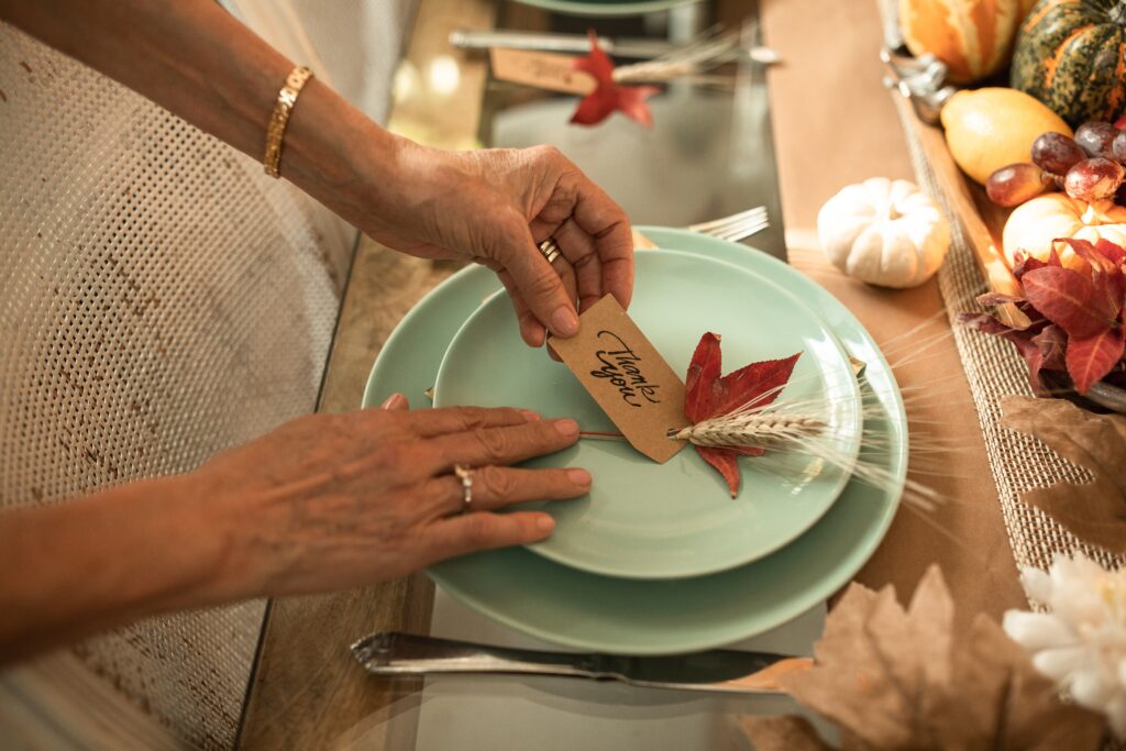 Image of hands adjusting a festive thanksgiving table setting with a notecard that reads "Thank You." This image is a decorative representation of the theme of this blog, "Is being grateful while you’re healing possible?"