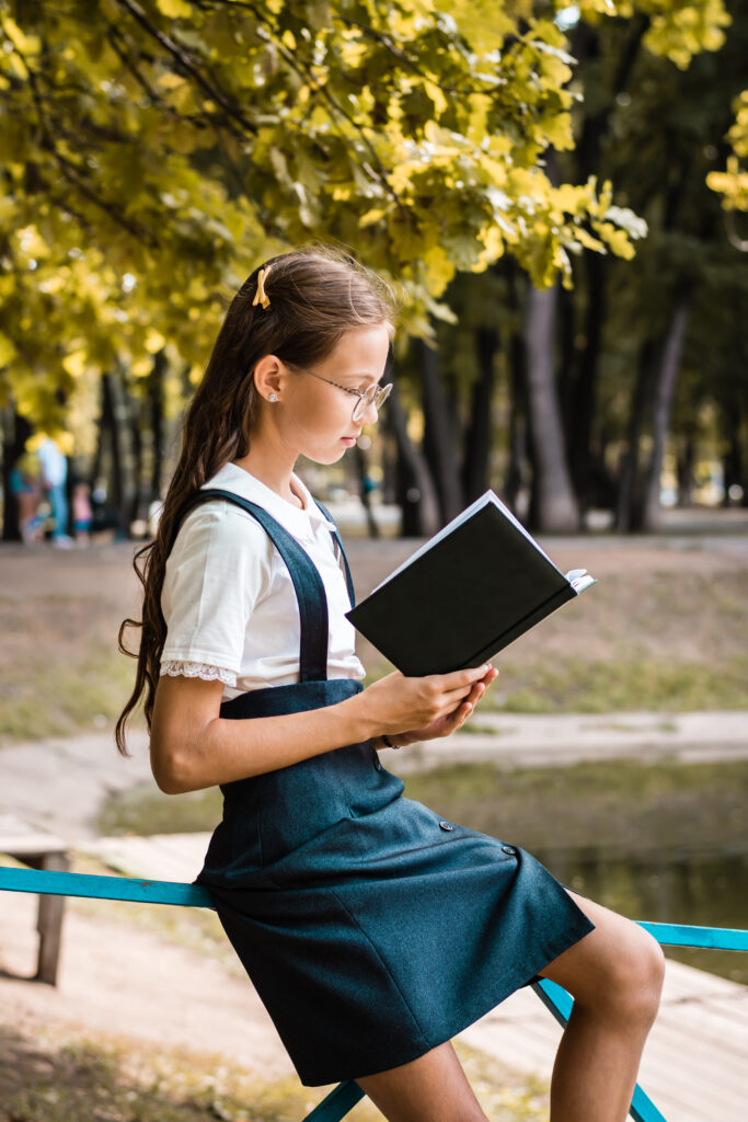 Teenage girl in school uniform reads a book on a warm day in the park. Vertical view