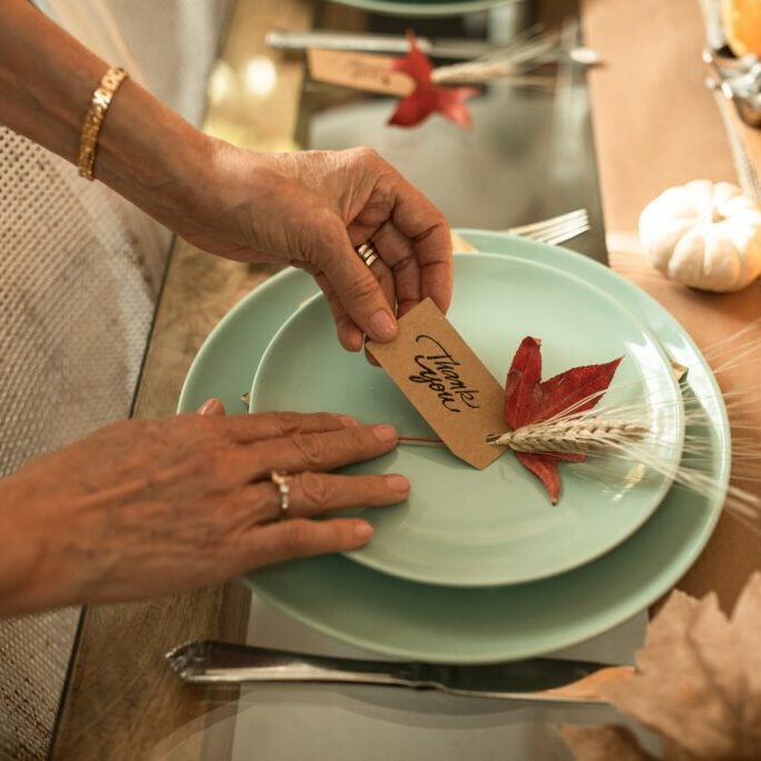 Image of hands adjusting a festive thanksgiving table setting with a notecard that reads "Thank You." This image is a decorative representation of the theme of this blog, "Is being grateful while you’re healing possible?"